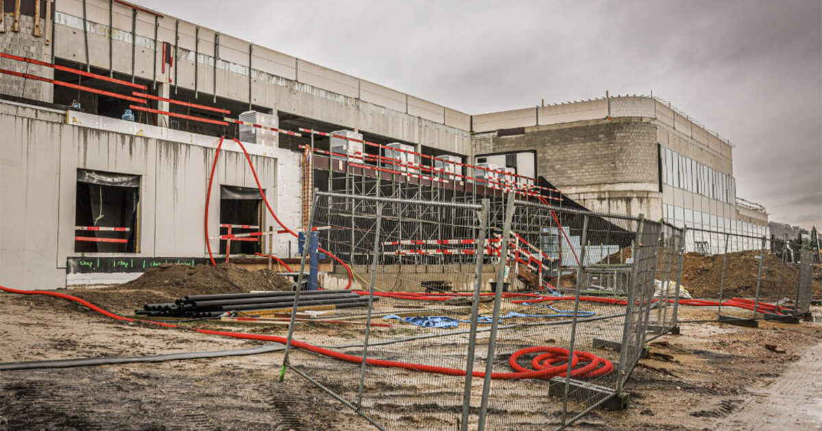 Bouwplaats met geraamte, steigers, blootliggende pijpen en bouwmaterialen verspreid op modderige grond onder bewolkte hemel.