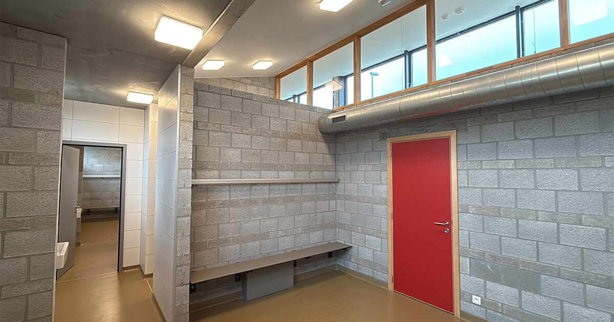 Een kamer met cementvrije bouwstenen muren, een rode deur, een metalen ventilatiekanaal, planken en hoge ramen; zichtbare aangrenzende kamer met betegelde muren en een gootsteen.