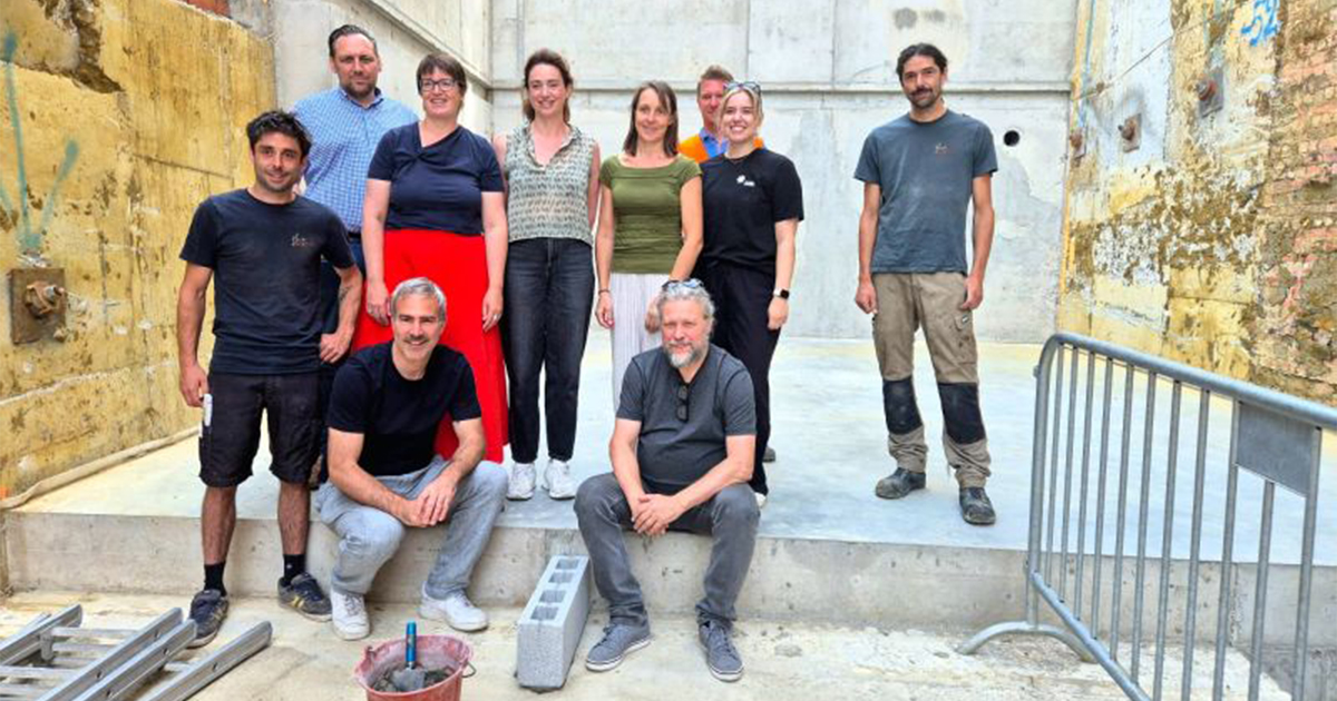 Tien mensen poseren samen op een bouwplaats; sommigen staan, anderen zitten op een betonnen richel. Op de voorgrond zijn bouwmaterialen en gereedschap te zien.