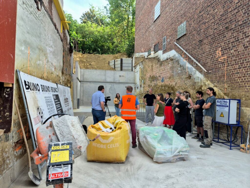 Een groep mensen staat op een bouwplaats naast bouwmaterialen; één persoon in een veiligheidsvest spreekt de groep toe bij een bakstenen muur en bouwmachines. De bouwplaats maakt deel uit van het CLT-project in Leuven, waar Masterwalls en prefabwanden worden toegepast als innovatieve bouwoplossingen.