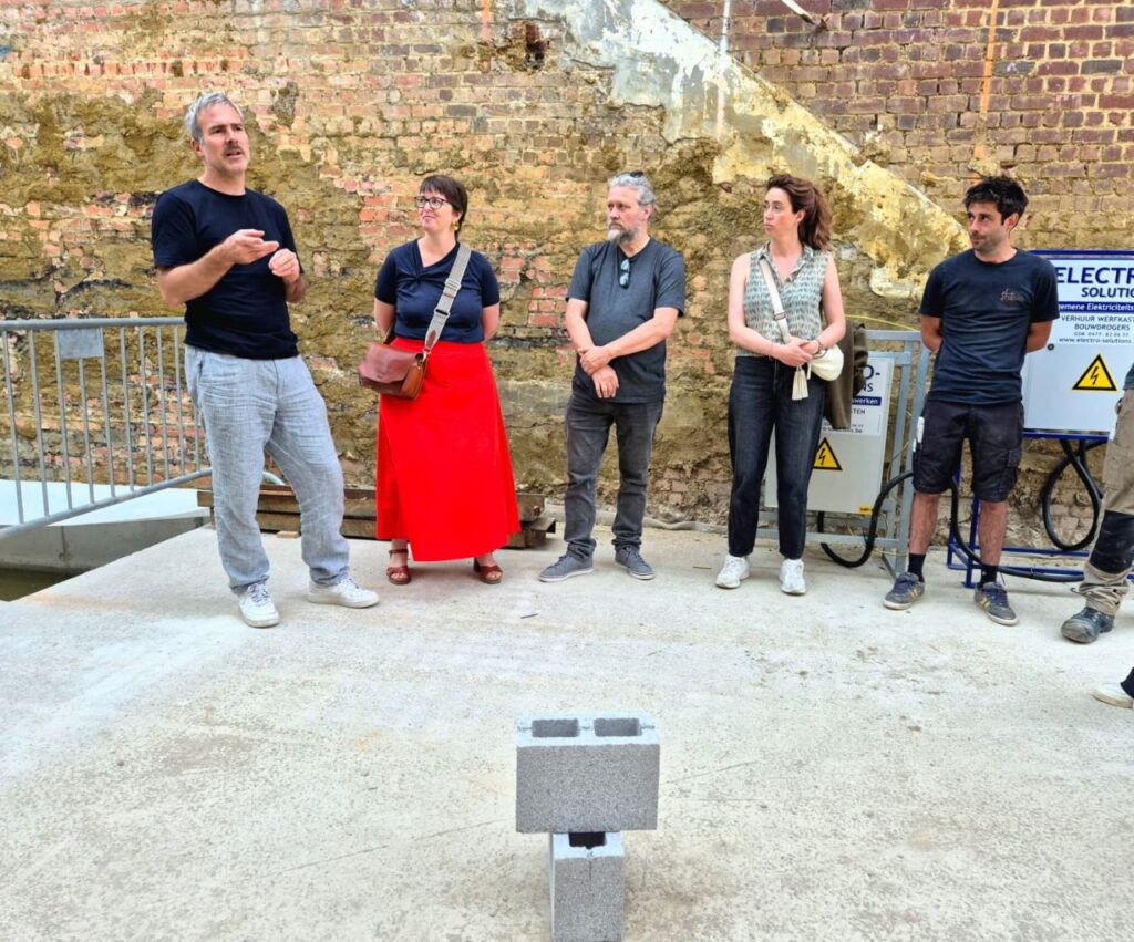 Vijf mensen staan op een betonnen ondergrond naast een sintelblok, met op de achtergrond een verweerde bakstenen muur en elektrische apparatuur — wellicht om de voortgang van het CLT-project in Leuven te bespreken en de prefabwanden van Masterwalls te bekijken in het kader van hun bouwplannen.