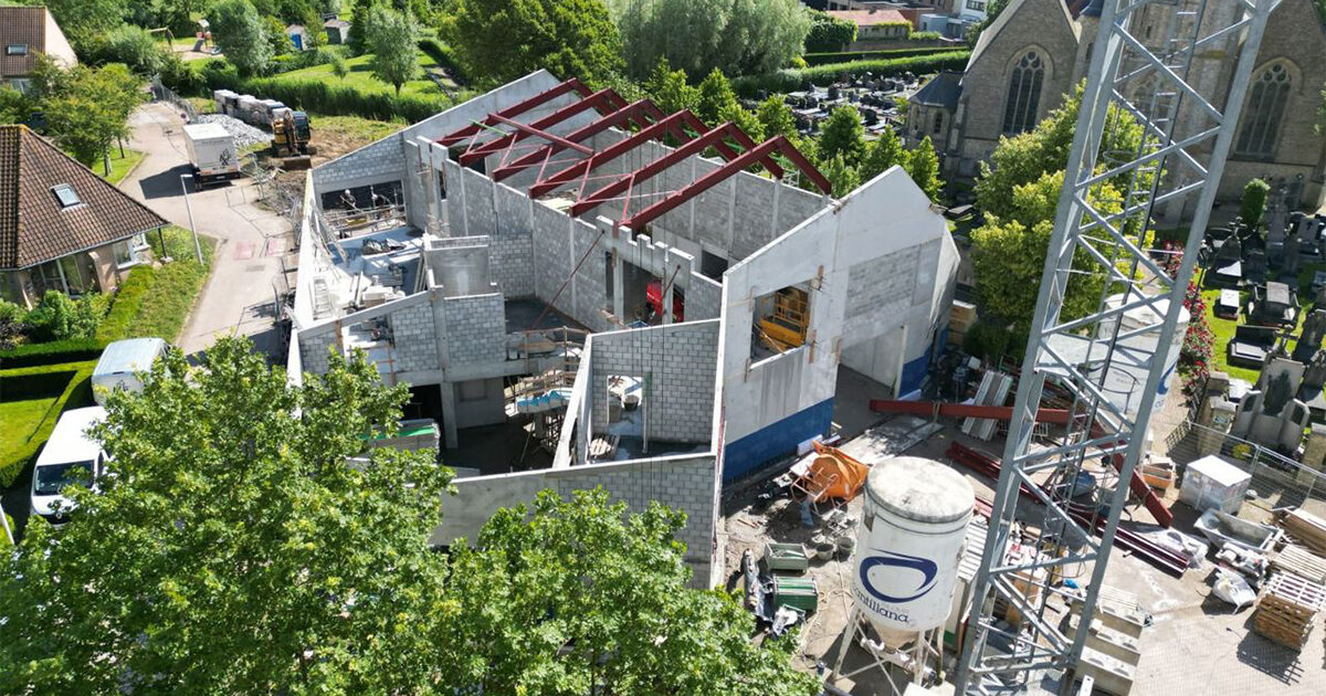 Luchtfoto van een gebouw in aanbouw, met gedeeltelijk voltooide muren gemaakt met Masterbloc, een alternatief voor beton. Blootliggende rode stalen balken, apparatuur en CO2-negatieve bouwsteenmaterialen zijn zichtbaar op de bouwplaats.
