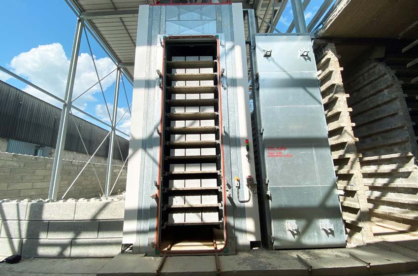 Een verticale industriële oven met gestapelde Carbstone cementvrije bouwsteen binnenin, gevestigd in een magazijn met stalen balken en gedeeltelijk zonlicht, die een koolstofnegatieve benadering van modern bouwen laat zien.