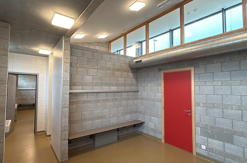 Een kamer met koolstofnegatieve muren van betonblokken, een rode deur, een bank, zichtbaar leidingwerk en bovenramen; een andere kamer met witte tegels is zichtbaar door een open deuropening. Een kamer met koolstofnegatieve muren van betonblokken, een rode deur, een bank, zichtbaar leidingwerk en bovenramen; een andere kamer met witte tegels is zichtbaar door een open deuropening.