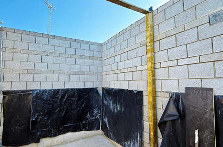 Gedeeltelijk geconstrueerd gebouw met cementvrije bouwstenen muren, zwarte waterdichtingsplaten langs de basis en een onafgewerkte houten balk onder een heldere hemel. Gedeeltelijk geconstrueerd gebouw met cementvrije bouwstenen muren, zwarte waterdichtingsplaten langs de basis en een onafgewerkte houten balk onder een heldere hemel.