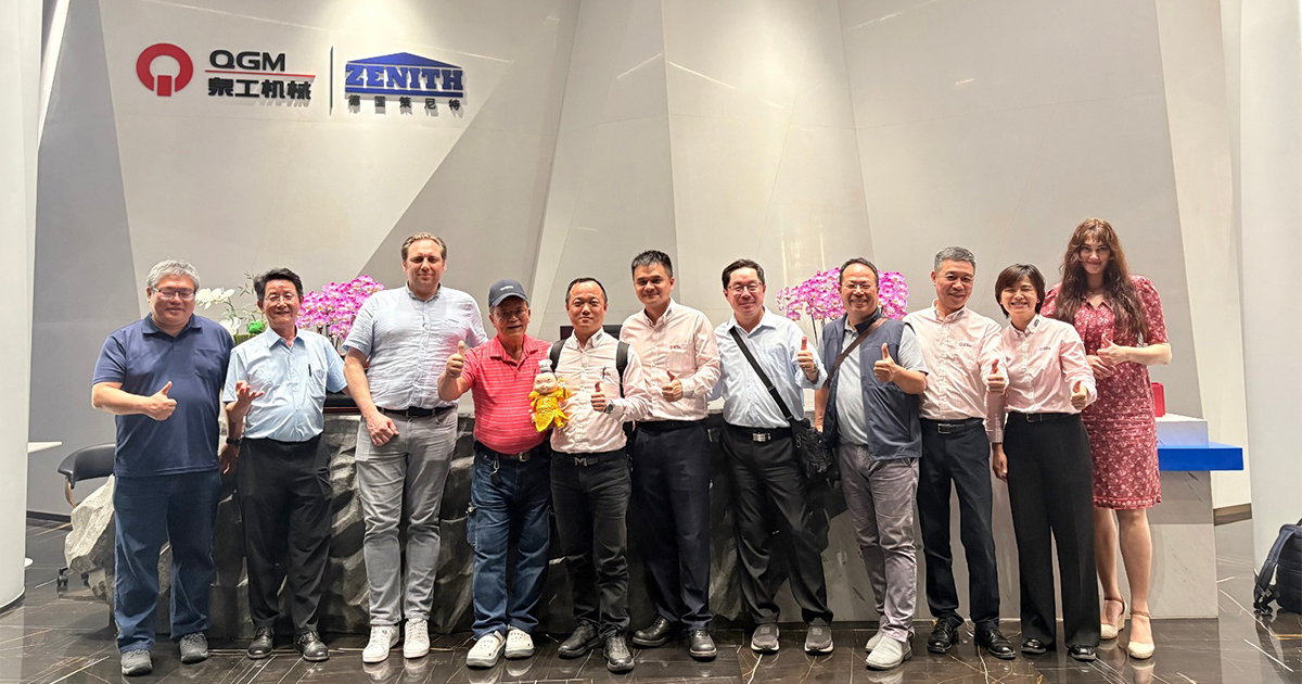 Un groupe de douze personnes se tient à l’intérieur devant les panneaux de QGM et ZENITH et pose pour une photo de groupe. Certaines personnes font un signe du pouce levé.